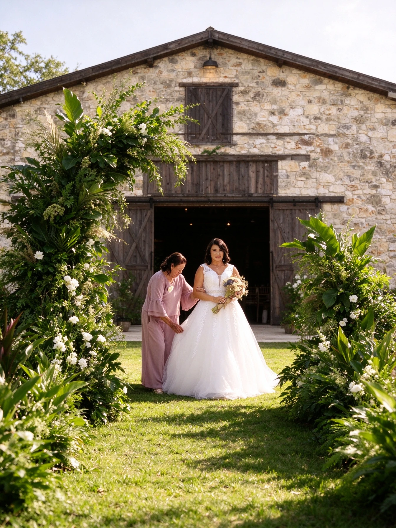 Mother adjusting the bride's gown near a stone barn and greenery arch
