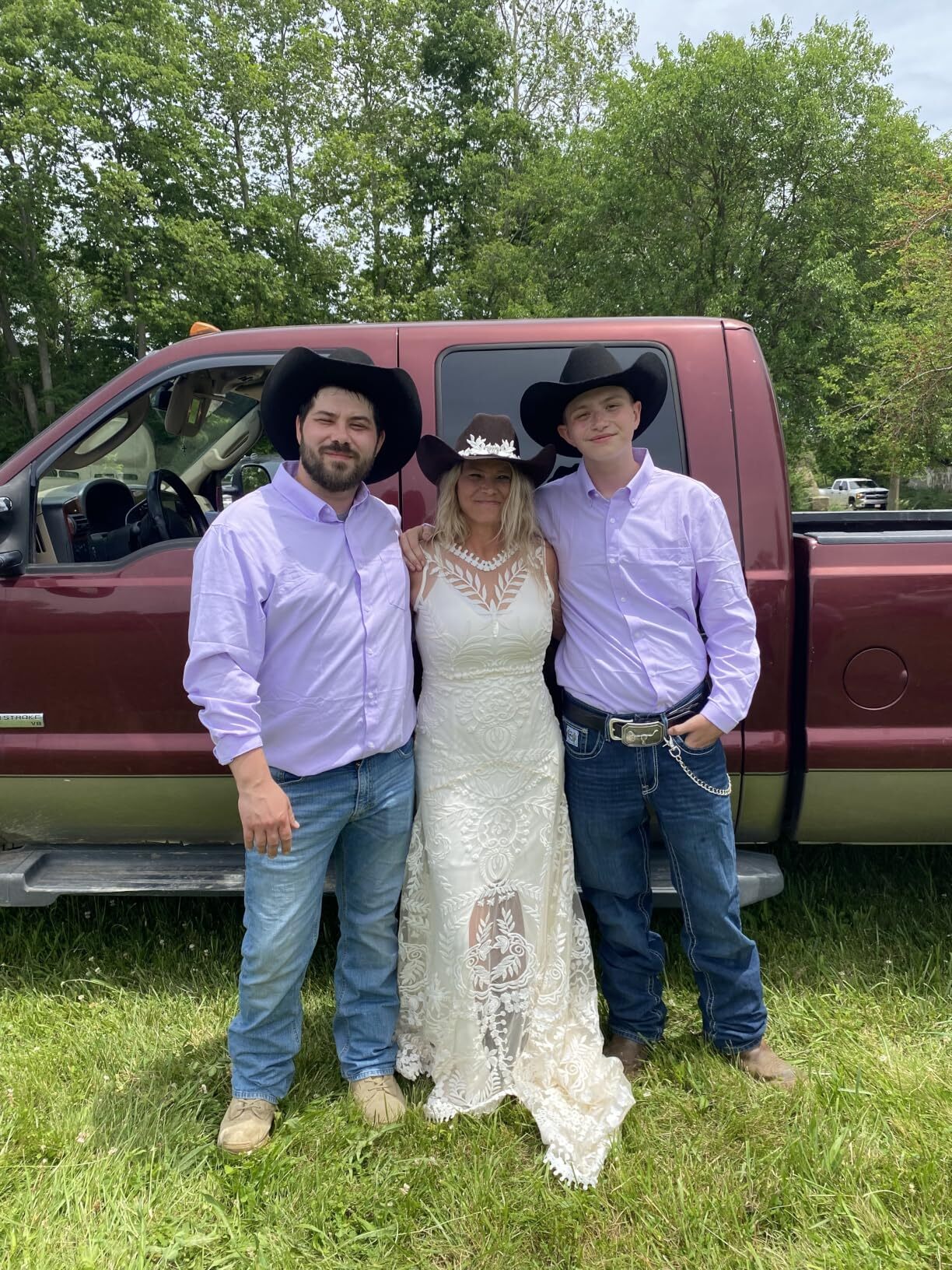 Bride wearing a sleeveless lace wedding dress with family beside a red pickup truck