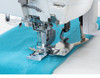 Top Cover Stitch in Action: A close-up action shot showing the machine actively stitching a top cover stitch on teal knit fabric. The needle plate, presser foot, and the built-in top cover hem guide are clearly visible, along with the L, M, R needle markings. Ideal for showcasing the signature top cover stitch feature.