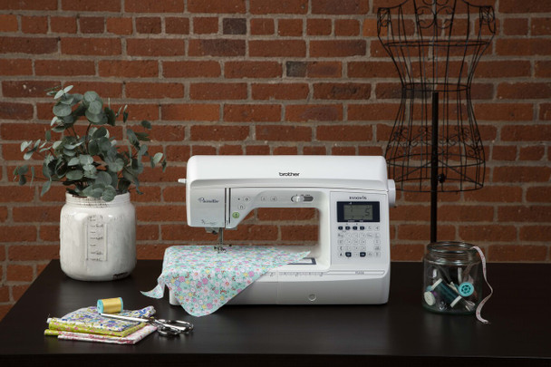Brother PS500 sewing machine on dark wooden table against brick wall with floral fabric, thread, and sewing accessories