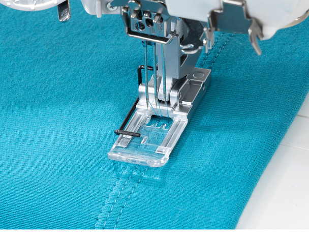 A tight macro shot of the clear view presser foot working on teal knit fabric, showing the needle plate, feed dogs, and a neatly finished cover stitch line. The transparent foot allows a clear view of stitch placement — ideal for highlighting this feature.