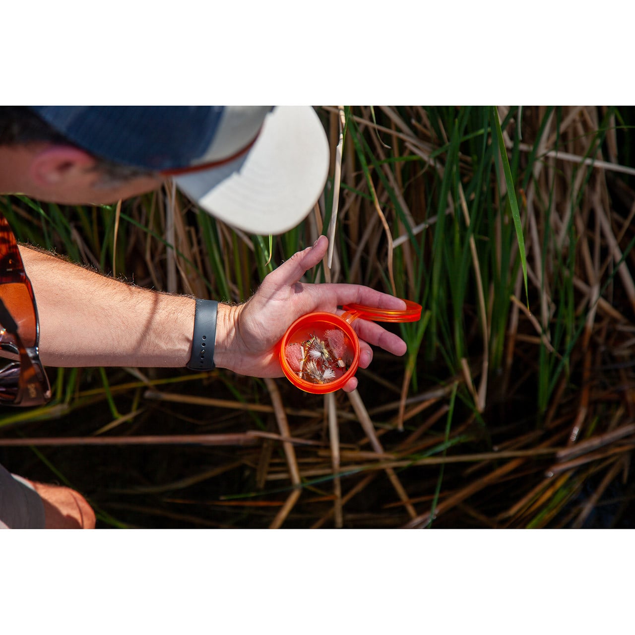 Fishpond Fly Puck with open lid and flies inside in an angler's hand beside a grassy streambank