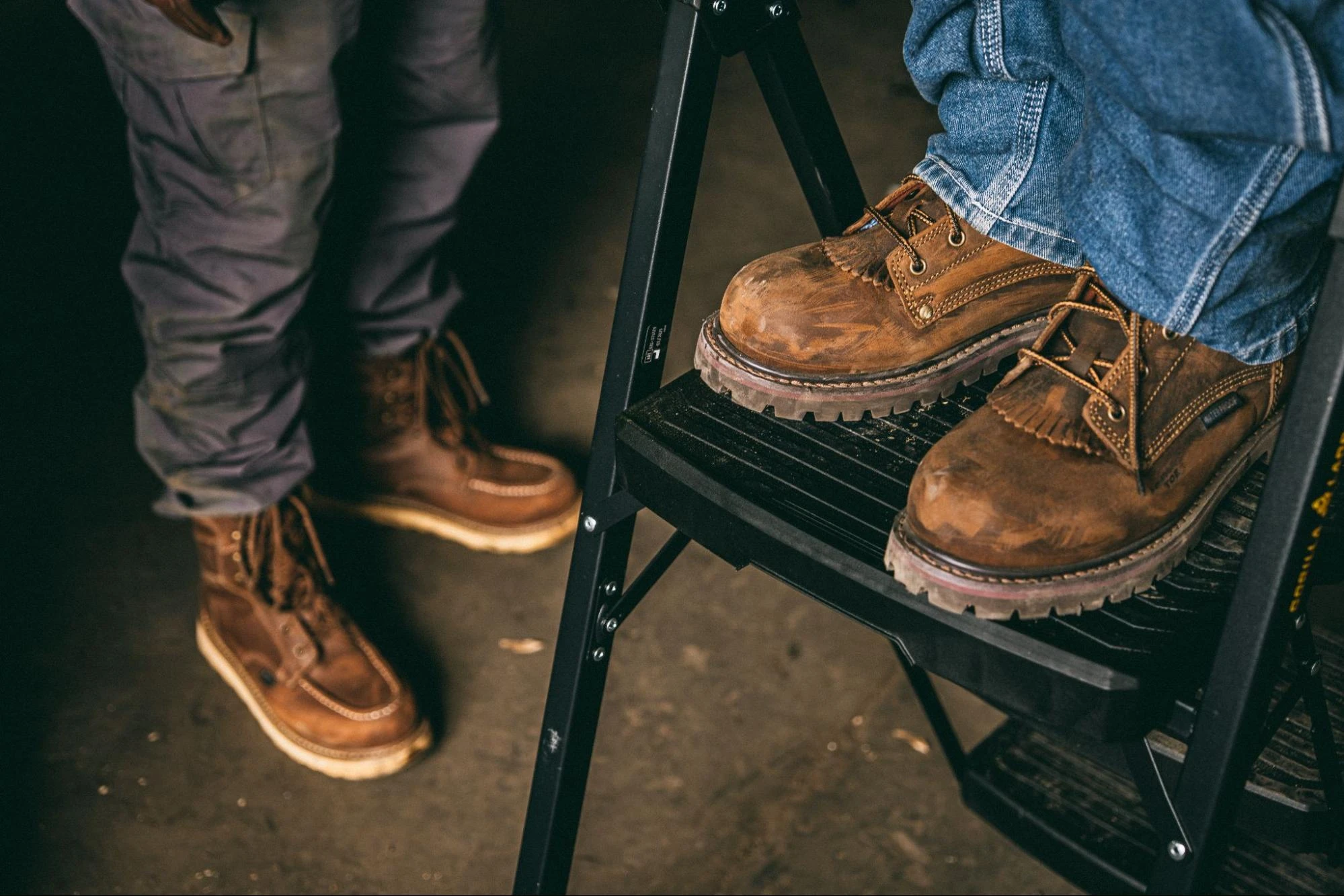 Two workers in brown Carolina work boots, one standing on a step ladder and one on the shop floor