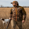 Man in Wildgrass camo jacket and hat with shotgun and dog in field