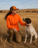 Woman in orange jacket and hat kneels with German Shorthaired Pointer in field.