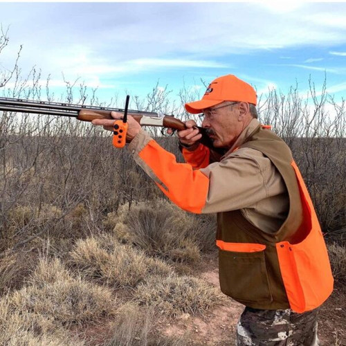 Man wearing orange vest and hat, aiming a shotgun in field.