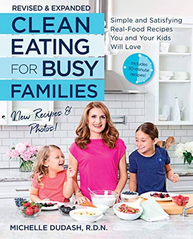 Front of Clean Eating for Busy Families with light blue and dark blue letters, with a background of a white kitchen, one lady in the middle and two children an either side of her, and food in bowls laid out in front of them on the front cover.