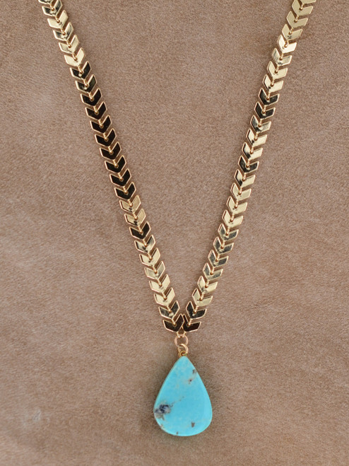 Genuine turquoise teardrop pendant on a polished gold arrow-link chain displayed on tan suede background.
