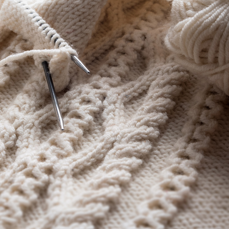 Knitting needles and a ball of cream yarn resting on an Aran cable-stitch panel