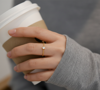 A close-up of a model's hand holding a coffee cup and wearing the Lone Spark Ring, with a single brilliant moissanite gemstone set in a thin, 18k gold-plated stainless steel.