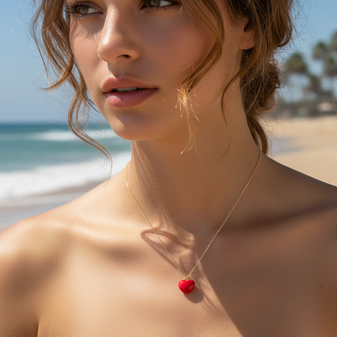 Close-up of a model at the beach, wearing a cherry red heart-shaped pendant on a delicate gold chain. The ocean with gentle waves is visible in the background.