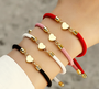 Hand wearing four Golden Heart Cord Bracelets by Keira K in white, pink, red, and black cords, featuring 18k gold-plated stainless steel heart charms. Delicate, trendy, and perfect for layering or any occasion.