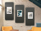 Three framed artworks mounted on dark acoustic panels against a white brick wall above lounge chairs.