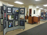 Library interior with wheeled divider panels displaying framed artwork, arranged as a gallery exhibit.