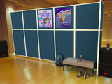 Blue freestanding partition wall with posters in a dance studio, with a bench, shoes, and a backpack on a wooden floor.