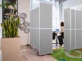 Gray freestanding partition panels forming a small workspace, with a person using a laptop beside indoor plants.