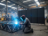 Welder kneeling and welding a steel frame inside an industrial workshop, with black wheeled divider panels behind him.