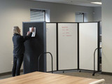 Person attaching a sheet to a black partition panel beside rolling whiteboard panels with handwritten notes in an office.