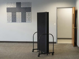 Folded black rolling privacy panels stored on a wheeled rack in an office hallway with wall art and an open door.