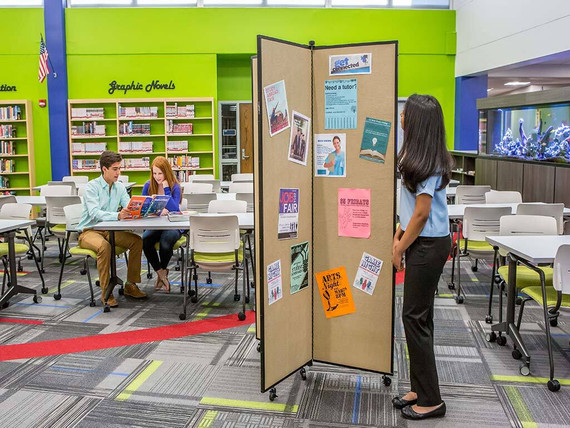 Student standing beside a wheeled bulletin board divider with flyers in a library classroom, while two students read at a table.