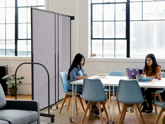 Small group working at a shared table beside a gray rolling partition panel in a bright office with large windows.
