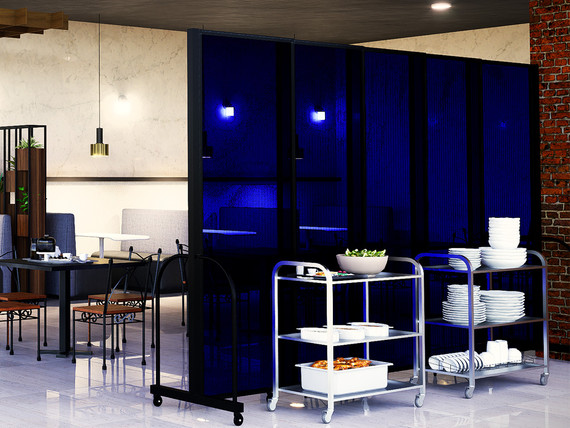 Restaurant interior with blue translucent partition panels on wheels, separating dining tables from serving carts stacked with plates.