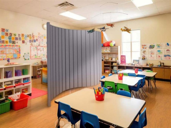 Preschool classroom with tables and colorful chairs, partially divided by a curved gray freestanding divider panel.