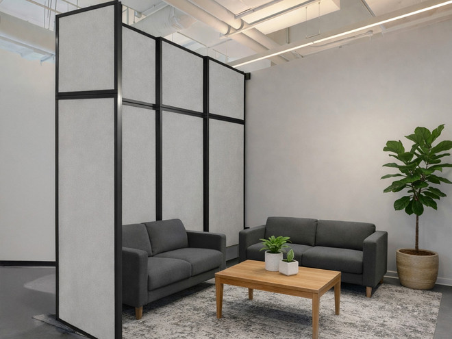 Modern office lounge with two dark gray sofas facing a wooden coffee table, separated by tall fabric-covered partitions, with a patterned rug and a potted plant.