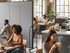 People working and socializing in a café-like workspace with tables, laptops, coffee cups, and a mobile fabric divider separating seating areas.