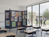 Classroom with desks and a wheeled divider covered in colorful posters, beside large windows showing students walking outside.