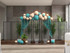 Balloon arch with teal and cream draping set in front of gray rolling partition panels, flanked by potted plants in a modern indoor space.