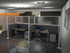 Two office desks enclosed by gray cubicle partition panels with computers, chairs, and storage cabinets in a modern workspace.