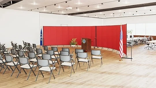 Two red room dividers form a backdrop behind a podium and rows of chairs at a rally.