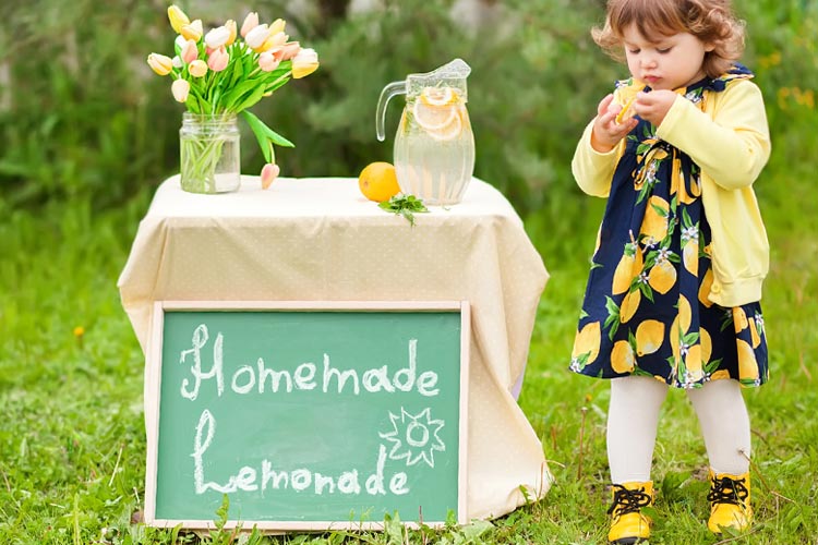 Cute toddler next to a lemonade stand outdoors for a carnival blog