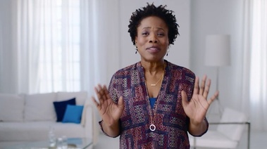 An elderly woman stands in her living room, hands raised, inviting and engaging with her surroundings