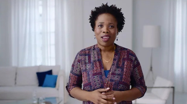 In a bright living room, a woman poses beside a modern white couch, surrounded by tasteful furnishings