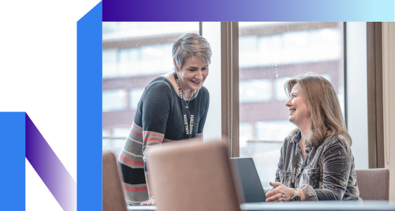 Two women are engaged in a conversation near a window. One is sitting with a laptop, and the other is standing, both smiling.
