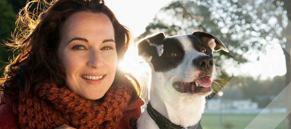 A woman wearing an orange scarf smiles next to a black and white dog outside on a sunny day