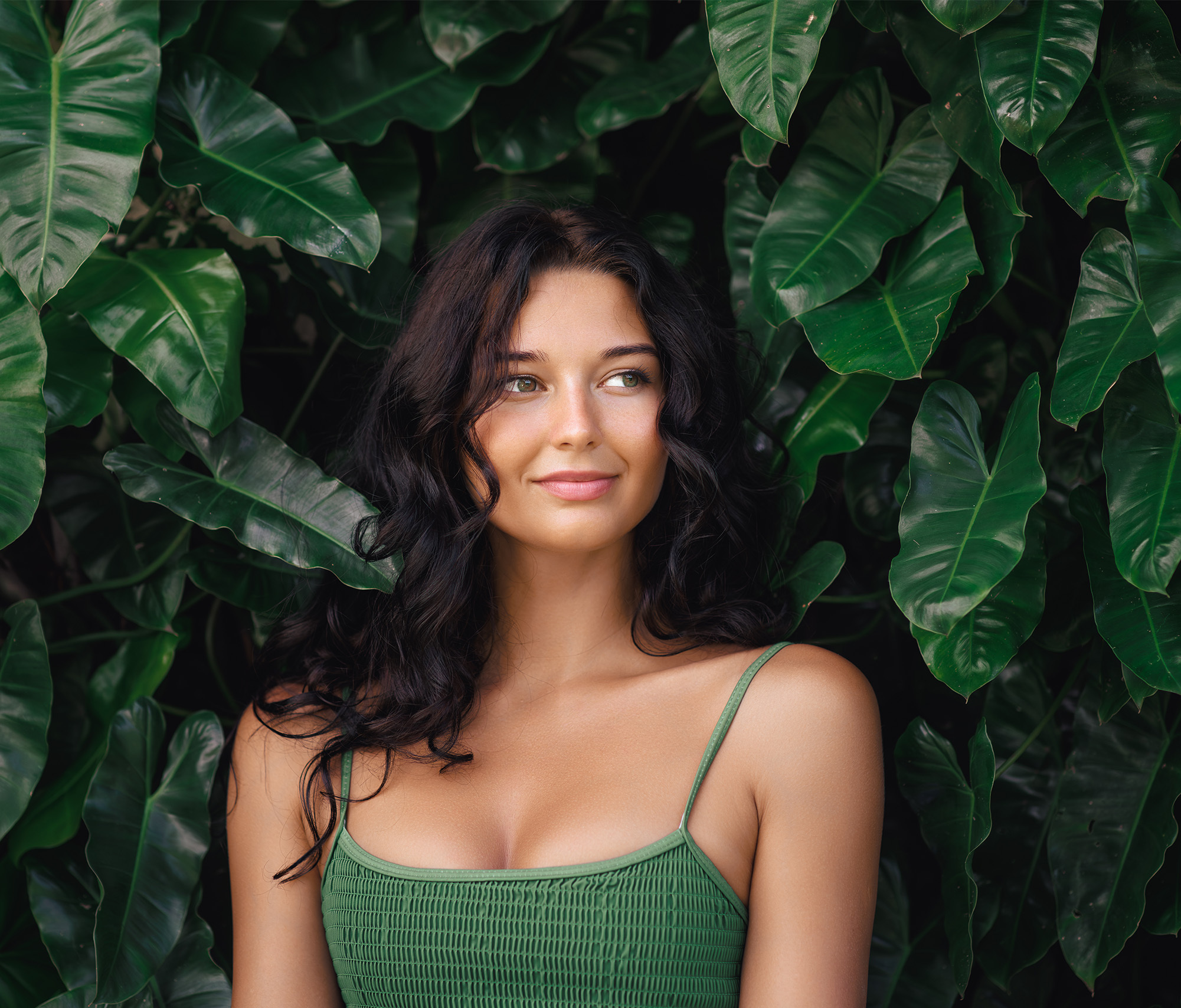 Woman smiling looking away from the camera with green foliage behind her.