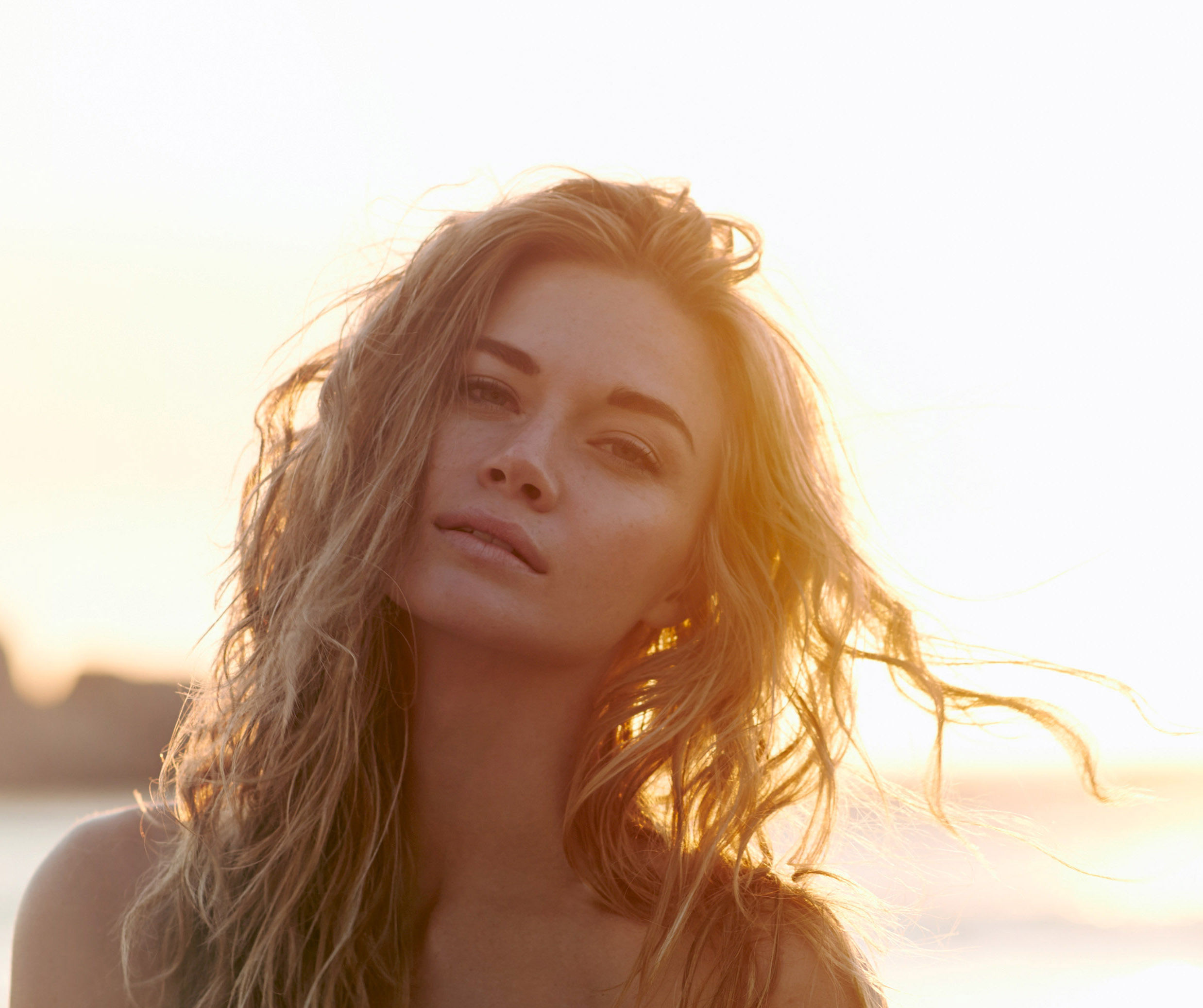 A woman with long, wavy hair photographed during golden hour with bright sunlight streaming from behind her. Her head is tilted slightly, and she has a serene, contemplative expression with her eyes partially closed. The backlighting creates a warm, glowing effect around her hair and silhouette. The background is softly blurred with warm, bright tones suggesting either sunrise or sunset. The overall mood is peaceful and ethereal.