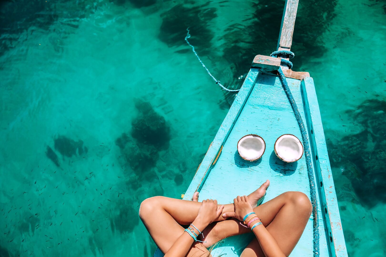 A person relaxing on the bow of a light blue wooden boat floating on crystal-clear turquoise water. They are lying with their legs crossed, wearing colorful ankle bracelets. Two halved coconuts sit on the boat's deck near their feet. The boat has weathered blue paint and rope rigging visible. The transparent water below reveals the sandy bottom and underwater vegetation, creating a perfect tropical paradise scene.