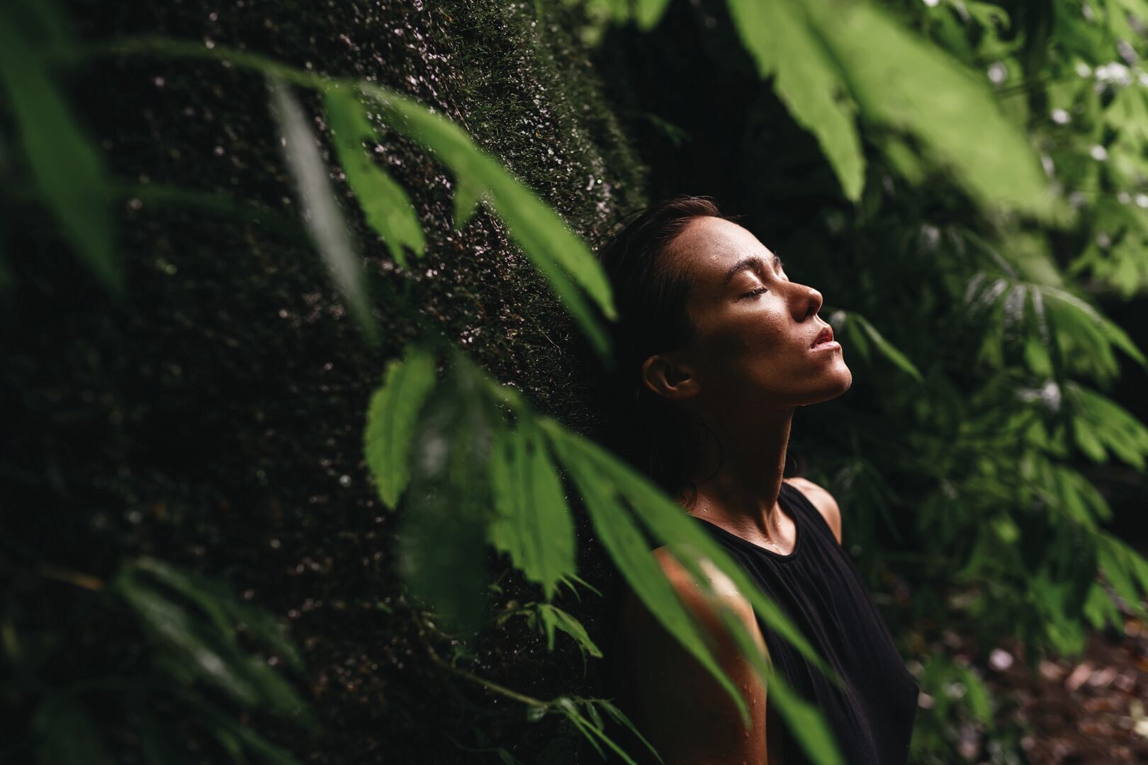 A woman with her eyes closed and face tilted upward in a peaceful, contemplative expression, surrounded by lush green tropical foliage. Water droplets sparkle in the air around her, creating a serene, nature-immersed atmosphere. She's wearing a dark-colored top and is enjoying a moment of tranquility in a rainforest.