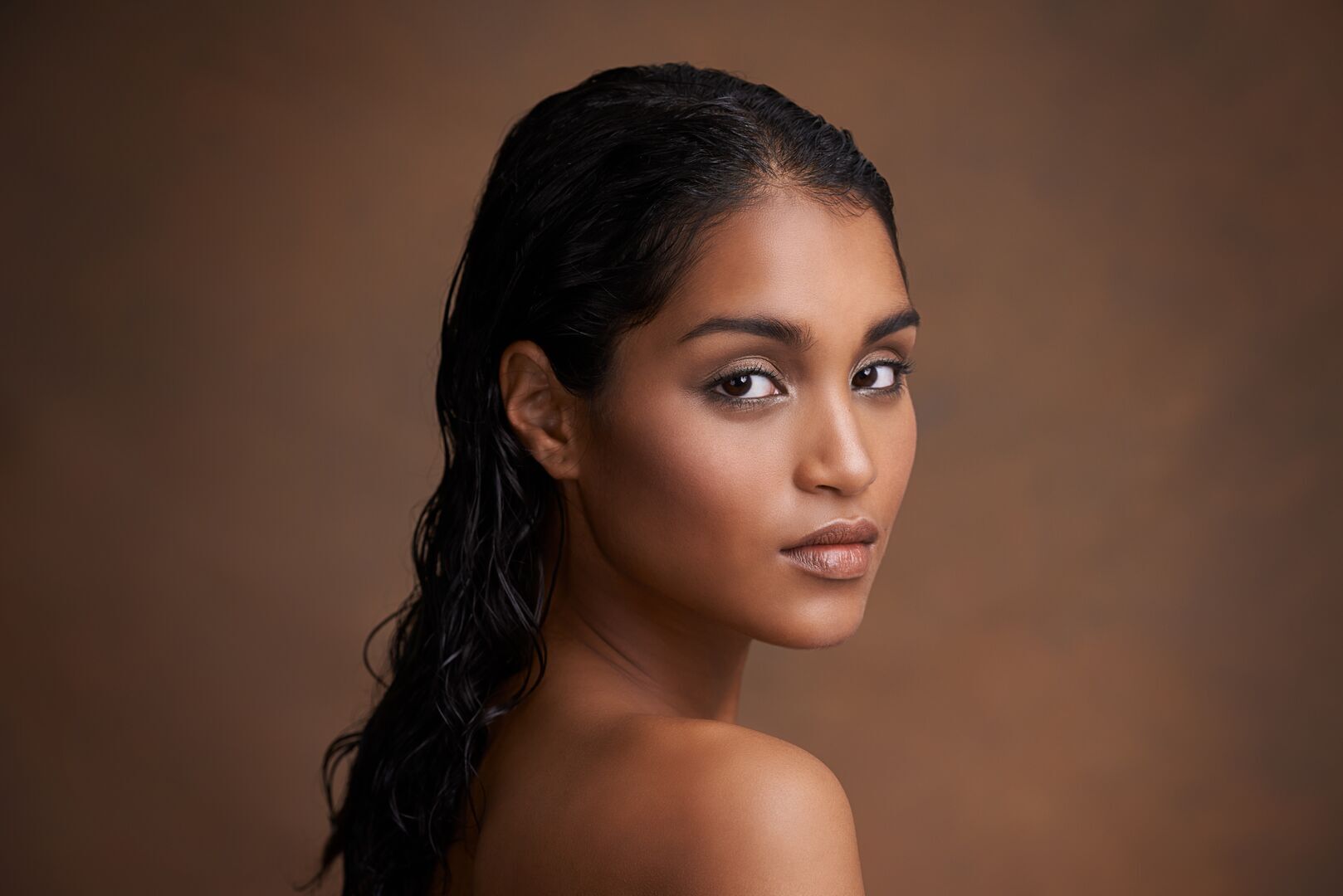 A portrait of a woman with dark hair slicked back, photographed from a three-quarter angle against a warm brown background. She has a confident, direct gaze looking slightly to the side, with natural makeup that emphasizes her eyes and lips. The lighting is soft and warm, creating gentle shadows that highlight her facial features. Her shoulders are bare, suggesting a minimalist portrait style. The overall composition has a professional, beauty photography aesthetic with rich, earthy tones.