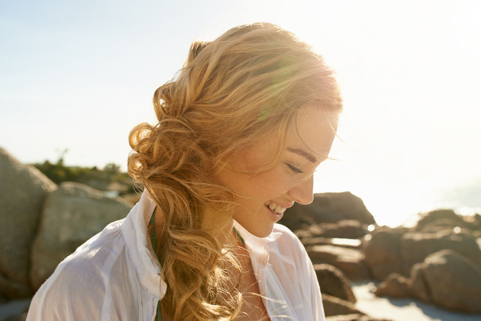 A woman with long, wavy blonde hair styled in a loose side braid, photographed in golden hour sunlight. She's wearing a white button-up shirt and looking down with a gentle smile. The warm sunlight creates a natural glow around her hair and face. In the background there are blurred stone structures or ruins, suggesting a coastal setting. The overall mood is bright and serene.