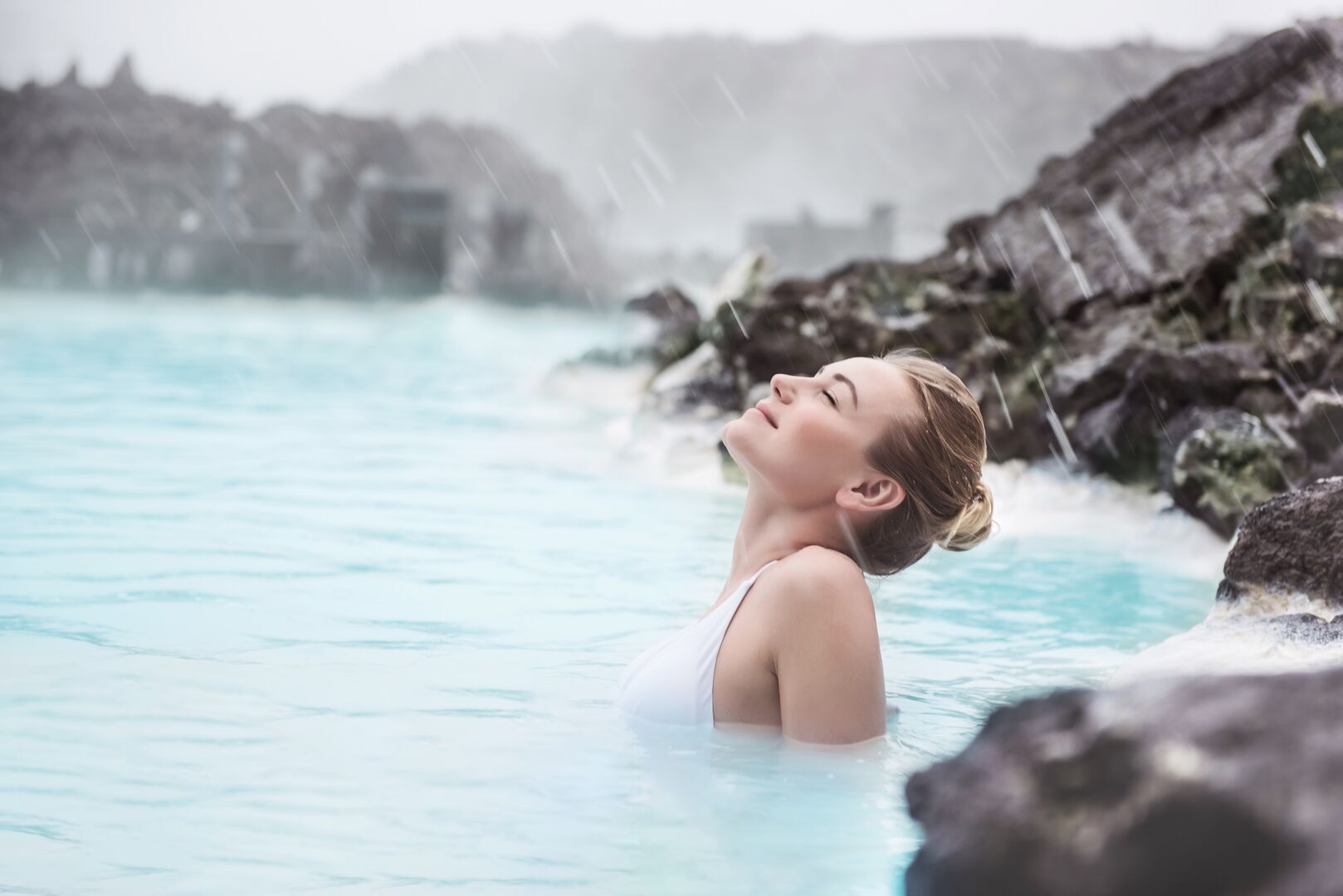 A woman with blonde hair pulled back relaxes in the famous milky blue waters of Iceland, her head tilted back with eyes closed in peaceful contentment. She is submerged up to her shoulders in the mineral-rich lagoon, surrounded by characteristic black volcanic rock formations. Steam rises from the warm waters, and the background shows Iceland's distinctive misty, mountainous landscape under an overcast sky. The scene captures the serene spa experience of Iceland's renowned hot springs.