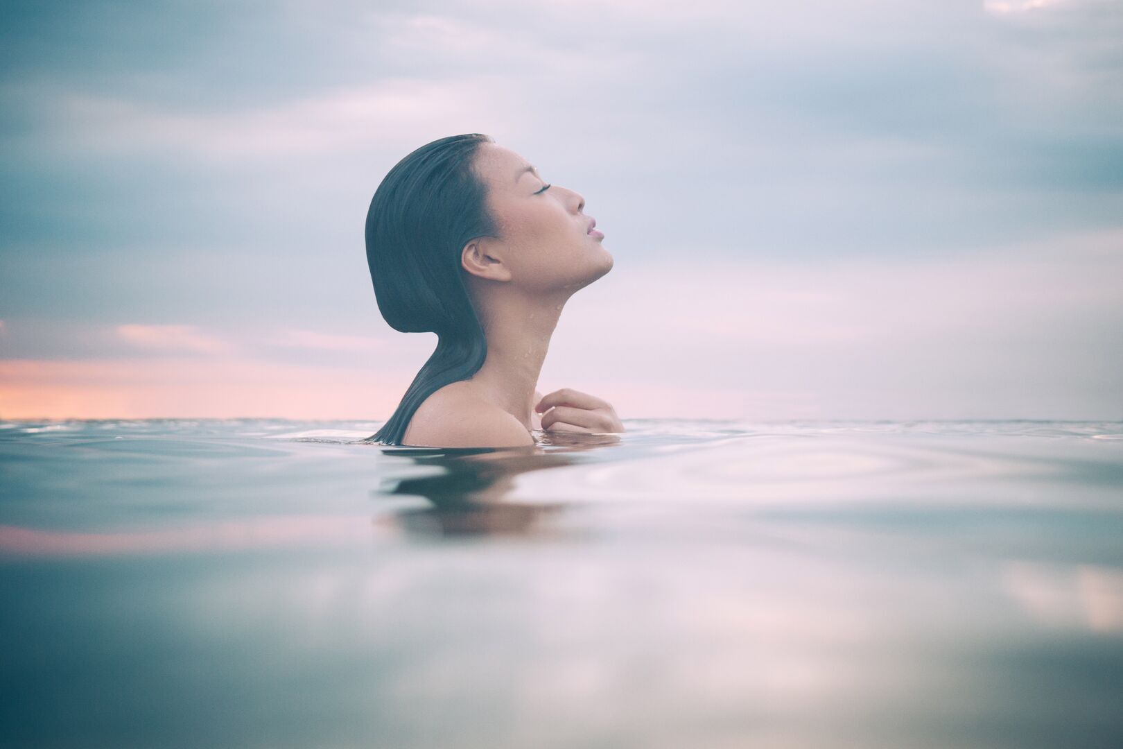 A woman with long dark hair floats peacefully in calm water, her head tilted back with eyes closed in a serene expression. She is partially submerged up to her shoulders, with soft ripples surrounding her. The background shows a dreamy, pastel-colored sky with pink and blue hues, suggesting either sunrise or sunset. The overall mood is tranquil and meditative.