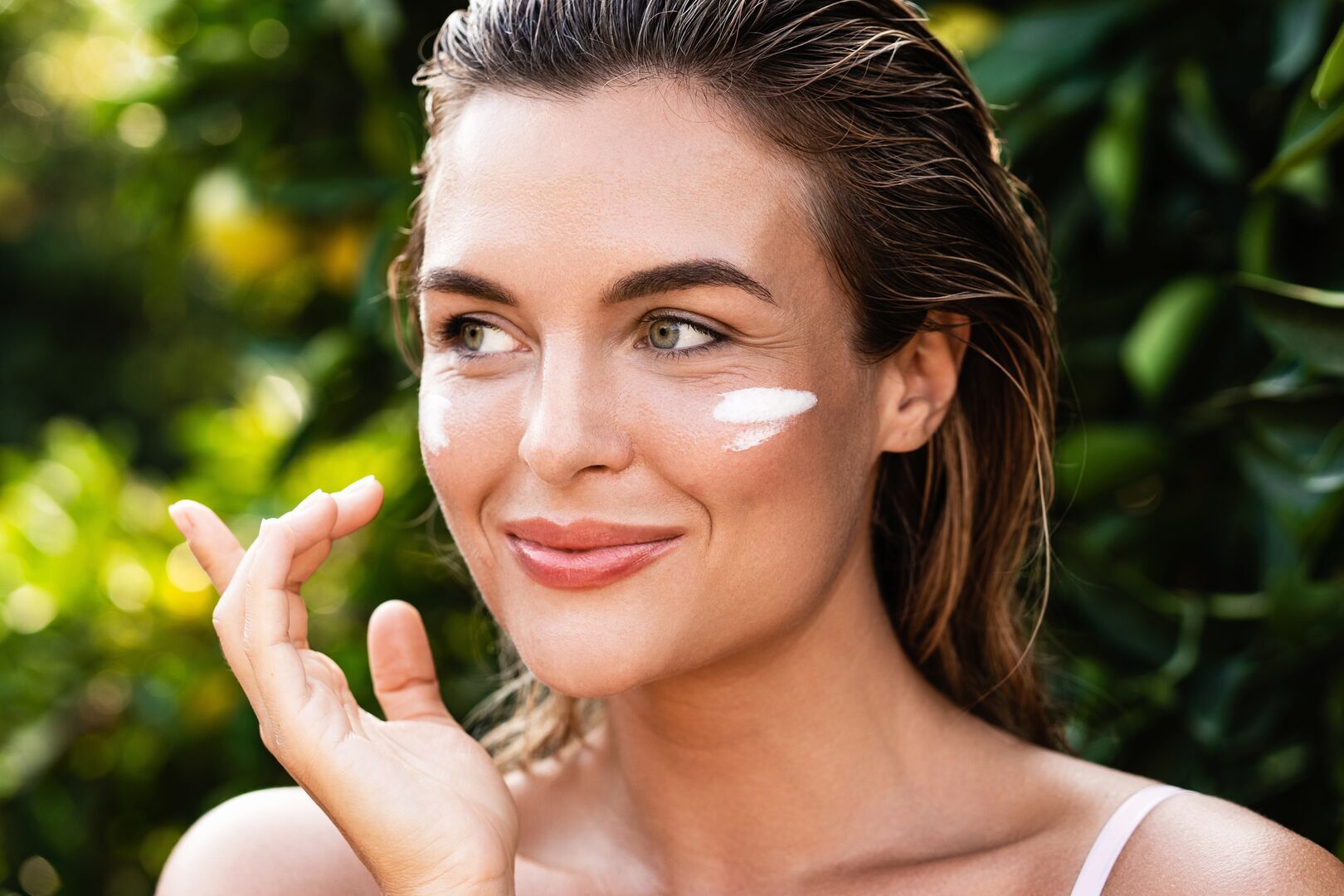 A woman with shoulder-length brown hair pulled back, smiling at the camera while applying Destination Patagonia Face Cream. She has a white streak of Destination Patagonia Face Cream on her right cheek and is holding her hand up near her face in a skincare application gesture. She's wearing a white top. The background shows a soft-focus natural setting with green foliage and warm, dappled sunlight, creating a fresh, outdoor skincare routine scene.