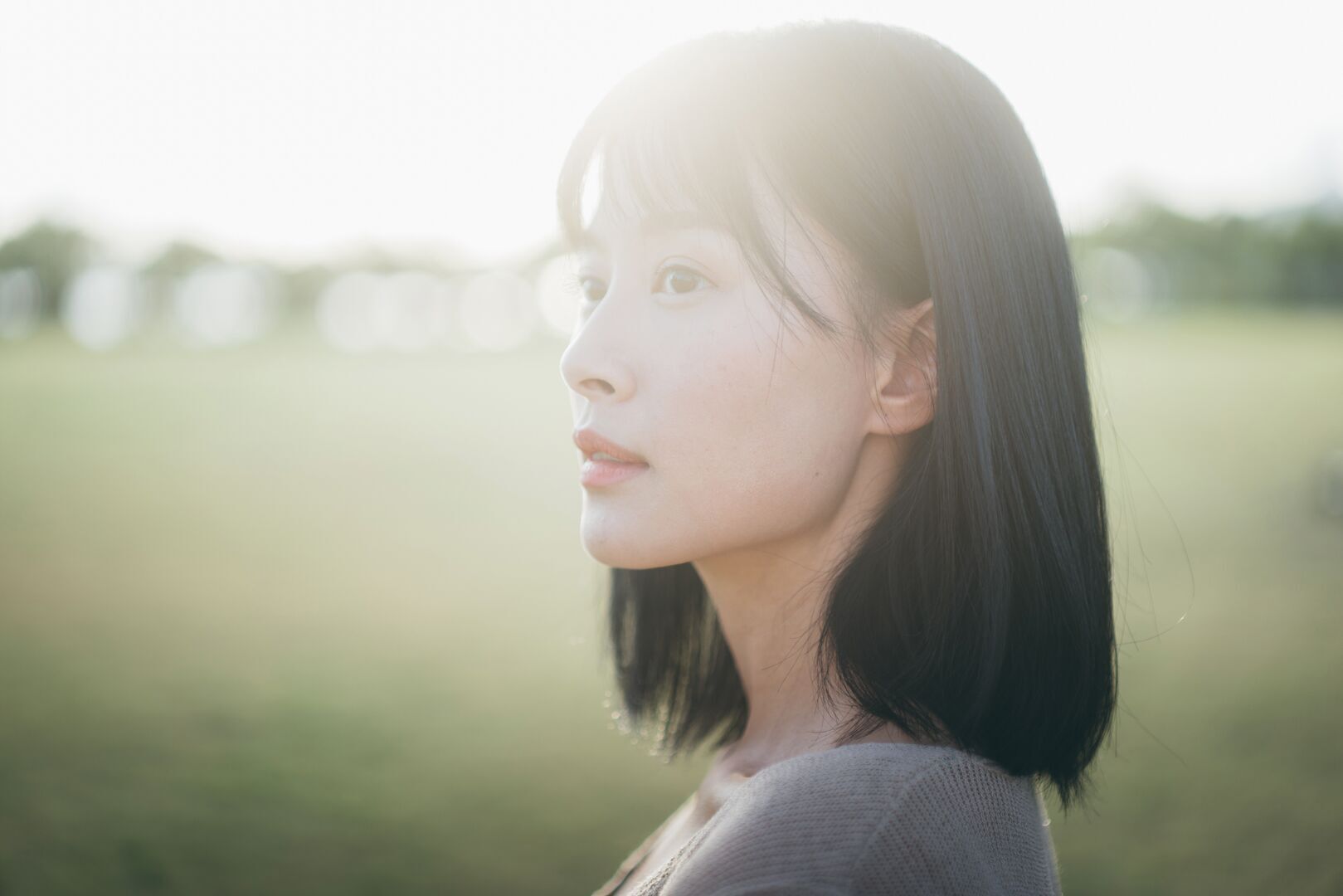 A young woman with shoulder-length dark hair in profile, gazing thoughtfully into the distance. She's wearing a light gray or beige sweater and is photographed outdoors with soft, warm lighting creating a gentle glow around her face. The background shows a blurred pastoral scene with green fields or meadows, giving the image a peaceful, contemplative mood.