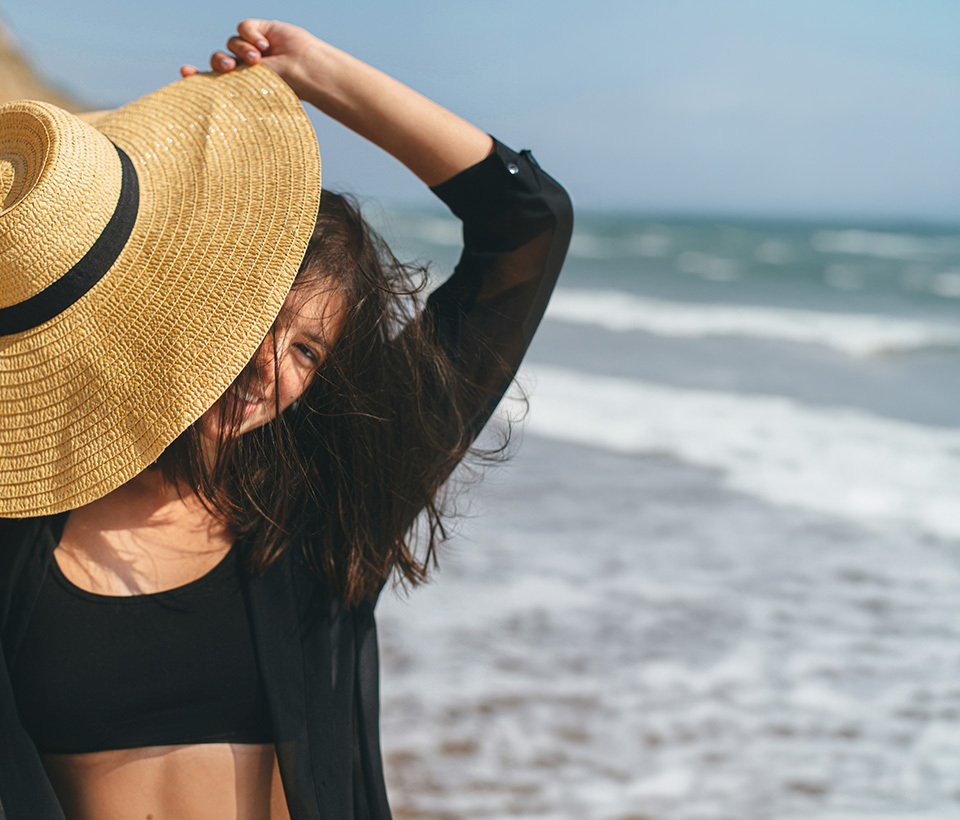 Woman smiling at the beach with a big floppy hat.
