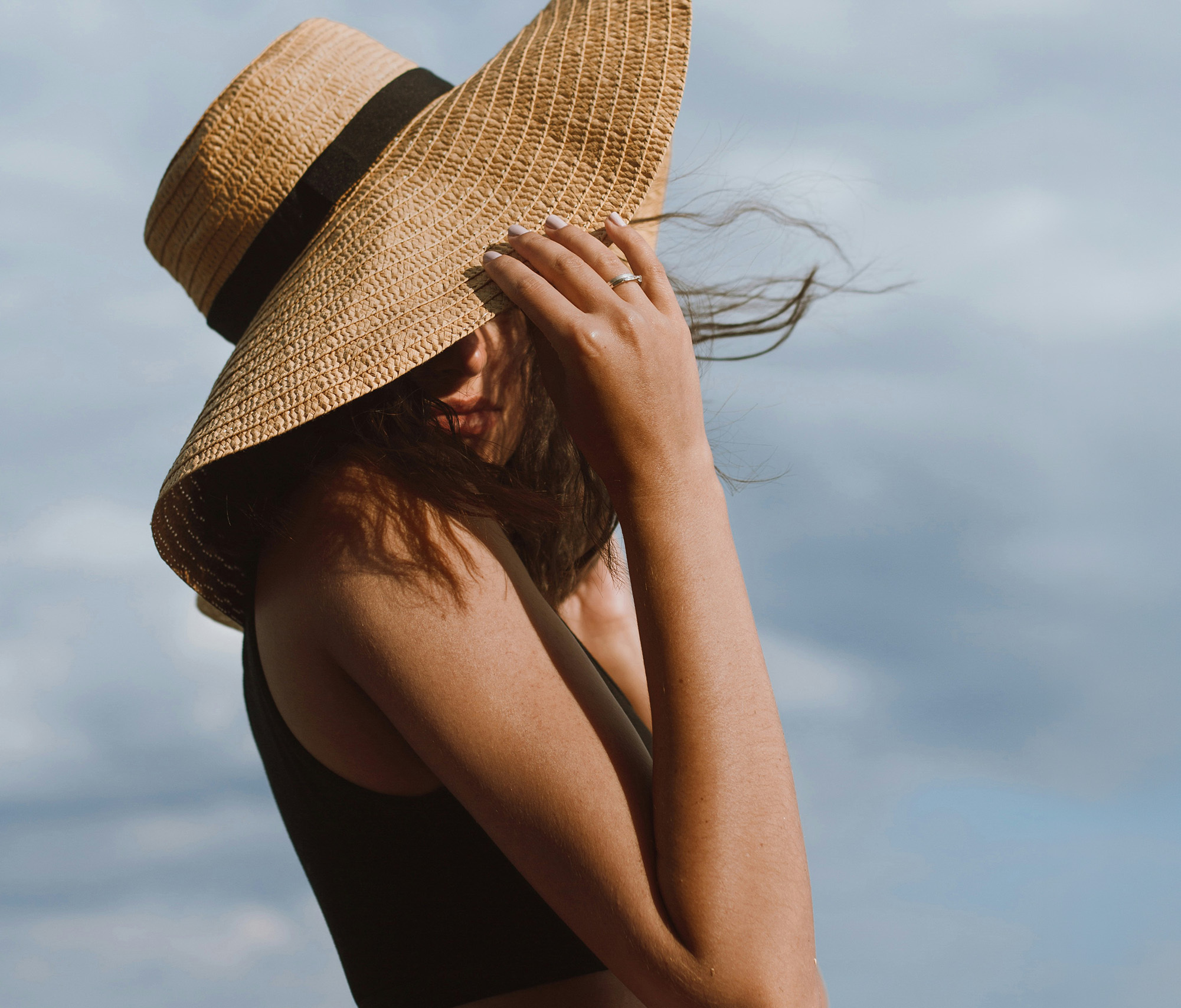 Woman wearing a big floppy straw hat, her hand gentle pulls the brim to cover her face.